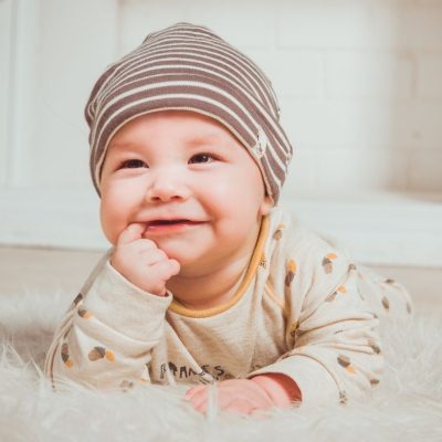 cute baby with finger in their mouth smiling with a beanie hat on their head