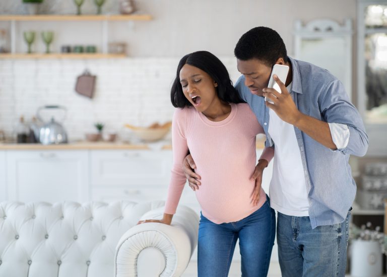 Pregnant African American woman having prenatal contractions, worried husband calling doctor on smartphone. Black lady giving birth to baby, her partner arranging medical help